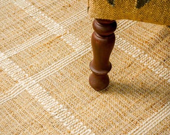 Könnte beinhalten: Ein gewebter Teppich mit einem strukturierten goldgelben und cremefarbenen Gittermuster. Ein einzelnes dunkelbraunes Holzmöbelbein mit gedrechseltem, rundem Design ruht auf der Oberfläche des Teppichs, der eine grobe, faserige Webstruktur aufweist.