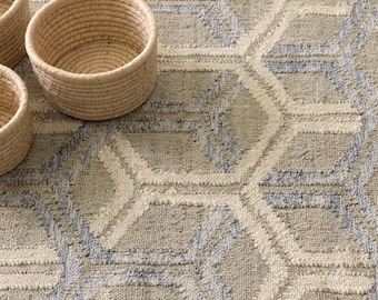 Könnte beinhalten: Ein gewebter Teppich mit einem sich wiederholenden geometrischen Sechseckmuster in Beige-, Creme- und sanften Blautönen, begleitet von drei runden, aus Naturfasern geflochtenen Körben auf der Teppichoberfläche.