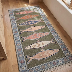 May include: A rectangular area rug featuring an olive green field with seven stylized fish in shades of pink, orange, blue, and cream. The rug has a blue border decorated with small floral motifs and finished with short fringe on both ends.