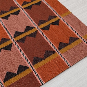 May include: A woven rug with a geometric pattern. The rug features alternating vertical stripes in shades of orange, brown, and pink. Black triangles and gold lines create a zig-zag design. The rug is made of a textured material.