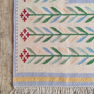 May include: A woven rug featuring a cream-coloured base with a repeating pattern of horizontal stems in green, blue, and peach, ending in red cross-stitch style flowers. The rug has a light purple border, a yellow stripe, and white fringe.