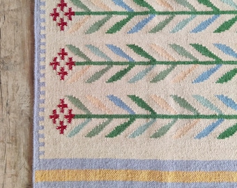 Könnte beinhalten: Ein gewebter Teppich mit cremefarbener Basis und einem wiederkehrenden Muster aus horizontalen grünen, blauen und pfirsichfarbenen Stielen, die in roten Kreuzstichblumen enden. Der Teppich hat einen hellvioletten Rand, einen gelben Streifen und weiße Fransen.