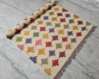 Könnte beinhalten: Ein rechteckiger Teppich mit cremefarbener Basis und einem wiederkehrenden Muster aus kleinen, kreuzförmigen Motiven in Rot, Gelb, Grün, Blau und Pink. Der Teppich ist an einem Ende teilweise aufgerollt und hat eine Fransenborte.