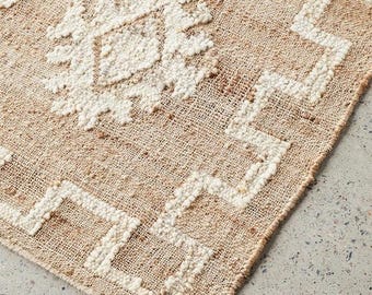 Könnte beinhalten: Ein gewebter Teppich mit einer Basis aus natürlichem, beigefarbenem Jute und erhabenen, weißen, getufteten geometrischen Mustern. Das Design umfasst sich wiederholende Rautenformen und Zickzack-Bordüren aus weichem, strukturiertem Garn.