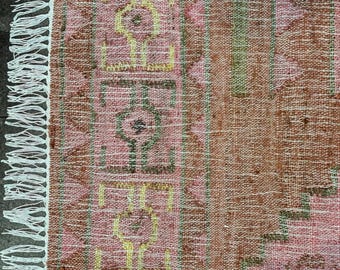Könnte beinhalten: Ein gewebter Textilteppich mit einer Farbpalette in Rosa und Braun sowie einem geometrischen Muster in Gelb und Grün. Der Teppich hat eine strukturierte Oberfläche und ist an der linken Kante mit weißen Fransen versehen, die einen Rand bilden.