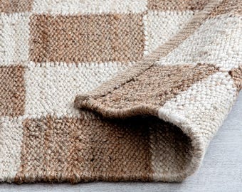 Könnte beinhalten: Ein gewebter Teppich mit einem sich wiederholenden Schachbrettmuster aus abwechselnden Quadraten in Naturbeige und Off-White. Der Teppich hat eine dicke, strukturierte Faserkonstruktion mit sichtbarem Gewebe und einer umgeschlagenen Ecke.