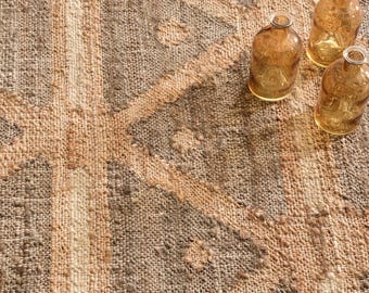 Könnte beinhalten: Ein gewebter Teppich mit einem geometrischen Muster aus beigen und braunen Dreiecken und einer strukturierten Oberfläche, begleitet von drei kleinen, klaren, bernsteinfarbenen Glasflaschen mit engem Hals und rundem Körper oben rechts.