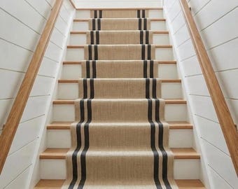 Könnte beinhalten: Eine Treppe mit einem beigen Jute-Läufer, der zwei vertikale schwarze Streifen in der Mitte aufweist. Die Treppenstufen bestehen aus hellem Holz mit weißen Setzstufen, flankiert von weißen, horizontal getäfelten Wänden und Holzhandläufen.