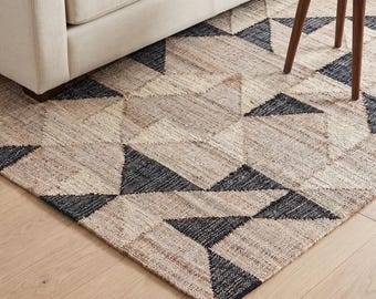 Könnte beinhalten: Ein rechteckiger Teppich mit einem geometrischen Muster aus Dreiecken in natürlichen Beige-, Hellbraun- und Schwarztönen. Die gewebte Struktur zeigt ein grobes, organisches Erscheinungsbild mit abwechselnden dunklen und hellen Dreiecksformen.