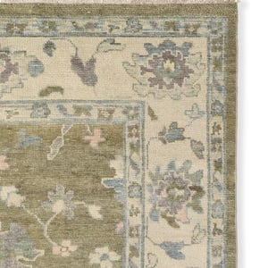 May include: A rectangular area rug featuring a central olive green field with a floral pattern in muted blue, pink, and cream. The rug has a wide cream border with a matching floral motif and a short, light-colored fringe along the top edge.