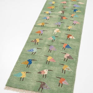 Op de afbeelding: Een rechthoekige loper in een zachte saliegroene tint, voorzien van een herhalend patroon van gestileerde vogels in diverse kleuren, waaronder blauw, oranje, geel, roze en paars. Het kleed is aan beide uiteinden afgewerkt met korte, crèmekleurige franjes.