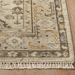 Puede incluir: Una alfombra rectangular con una base de color crema, textura tejida y un borde decorativo. El patrón incluye motivos geométricos y florales complejos en tonos marrón oscuro, oro apagado y azul claro, rematada con flecos en el borde.