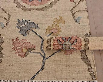 Könnte beinhalten: Ein beigefarbener Teppich mit einem sich wiederholenden Blumenmuster in Rosa-, Blau- und Brauntönen. Der Teppich hat ein strukturiertes Gewebe und Fransen an der Unterkante. Ein Teil des Teppichs ist umgerollt und zeigt dasselbe Blumenmuster.