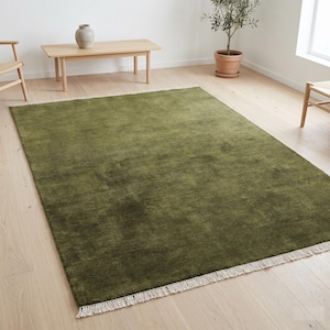 May include: A large, rectangular olive green rug with a textured surface and a fringe border. The rug is placed on a light-colored wooden floor in a room with natural light. A small wooden table and chairs are visible in the background.