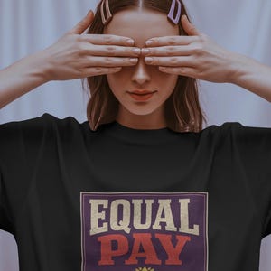 Può includere: T-shirt nero con la scritta "EQUAL PAY" in stile vintage, con la scritta "AAPI WOMEN DESERVE IT ALL" sotto. La maglietta è indossata da una persona con i capelli castani e due fermagli. La persona si copre gli occhi con le mani.