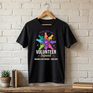 Peut inclure: T-shirt noir avec un graphisme coloré de mains formant un cercle autour d'un cœur. Le texte indique "TOGETHER WE" au-dessus et "VOLUNTEER Squad" en dessous, avec "MAKING A DIFFERENCE - TOGETHER" en bas.