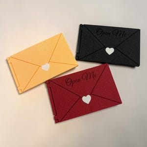 Puede incluir: Tres cajas decorativas con forma de sobre en amarillo, negro y rojo. Cada caja presenta un corazón blanco y las palabras "Open Me". Las cajas están diseñadas para parecer sobres.