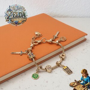 Pode incluir: Pulseira dourada com berloques temáticos, incluindo espadas, escudos e um emblema verde. A pulseira é exibida em um caderno laranja com o texto "The Legend of Zelda Breath of the Wild" no canto superior esquerdo.