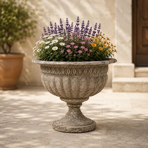 Puede incluir: Una gran urna de piedra desgastada llena de una variedad de flores coloridas. Las flores incluyen margaritas blancas, lavanda púrpura, flores rosas y amarillas. La urna tiene un diseño ornamentado y se asienta sobre una base de pedestal.