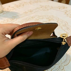 May include: Black handbag with brown leather accents. The bag has a top flap with a snap closure and a gold-colored zipper. The interior is black, and the bag appears to be made of nylon.