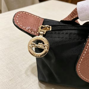 May include: Black handbag with a gold-tone zipper and a leather handle. The bag features a gold-tone charm with the brand name and the year 1948. The handle is a light brown color, and the bag is made of a durable material.