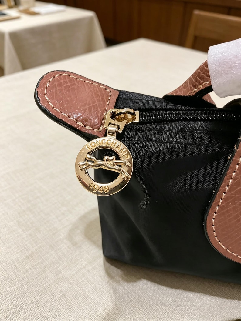 May include: Black handbag with a gold-tone zipper and a leather handle. The bag features a gold-tone charm with the brand name and the year 1948. The handle is a light brown color, and the bag is made of a durable material.