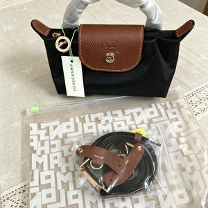 May include: Black handbag with a brown leather flap and handles. The bag has a gold-colored clasp and a tag that says "LONGCHAMP". A separate shoulder strap is included.