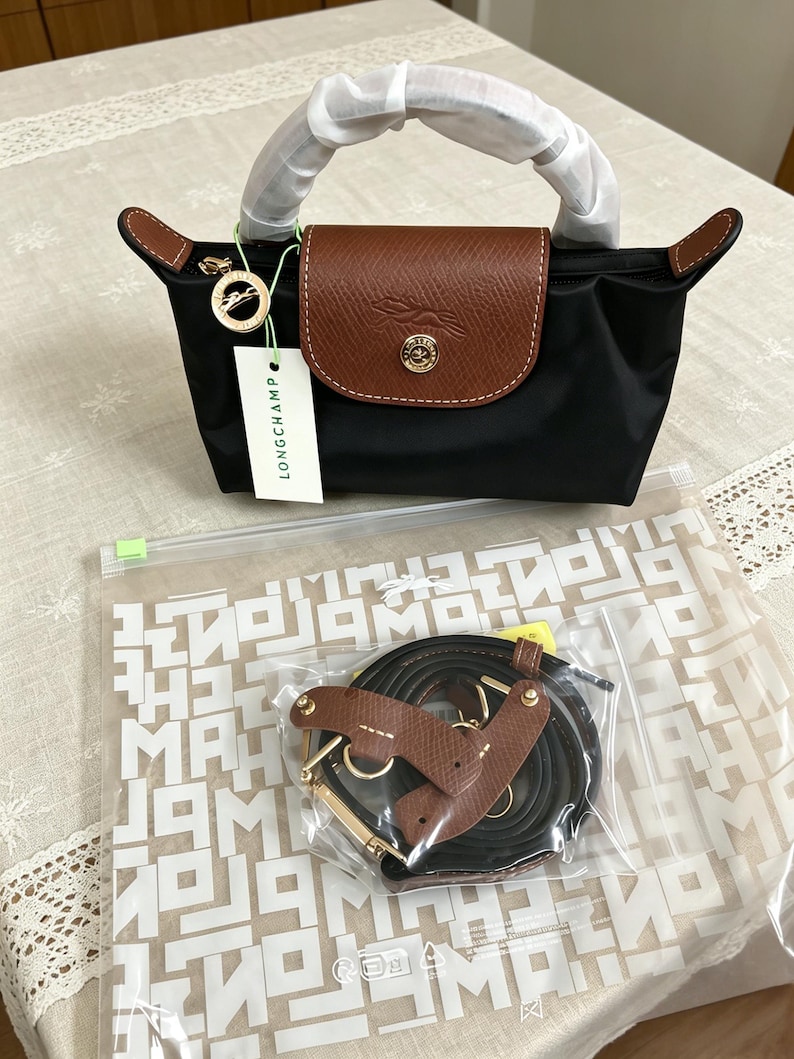 May include: Black handbag with a brown leather flap and handles. The bag has a gold-colored clasp and a tag that says "LONGCHAMP". A separate shoulder strap is included.