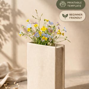 Könnte beinhalten: Eine beige, buchförmige Vase gefüllt mit bunten Wildblumen. Die Vase hat ein strukturiertes, vertikal gestreiftes Design, und der Text "BOOK VASE PRINT & MAKE" steht oben. Zusätzlicher Text lautet "Printable Template" und "Beginner Friendly".