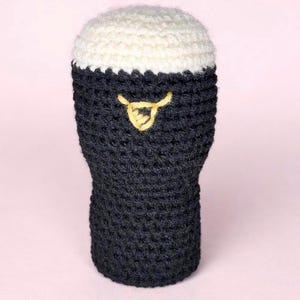 May include: A crocheted beer glass with a dark gray body and a white, frothy top. A yellow, decorative element is stitched on the front. The handmade item is likely a decorative piece or a cozy.