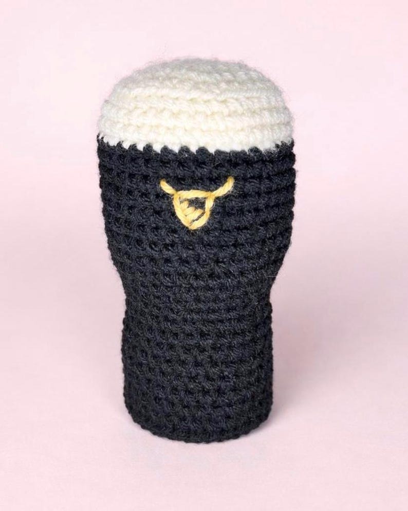 May include: A crocheted beer glass with a dark gray body and a white, frothy top. A yellow, decorative element is stitched on the front. The handmade item is likely a decorative piece or a cozy.