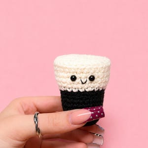 May include: A small, crocheted drink with a smiling face. The top is cream-colored, the middle is black, and the bottom is a dark burgundy with small white dots. The background is a solid pink.