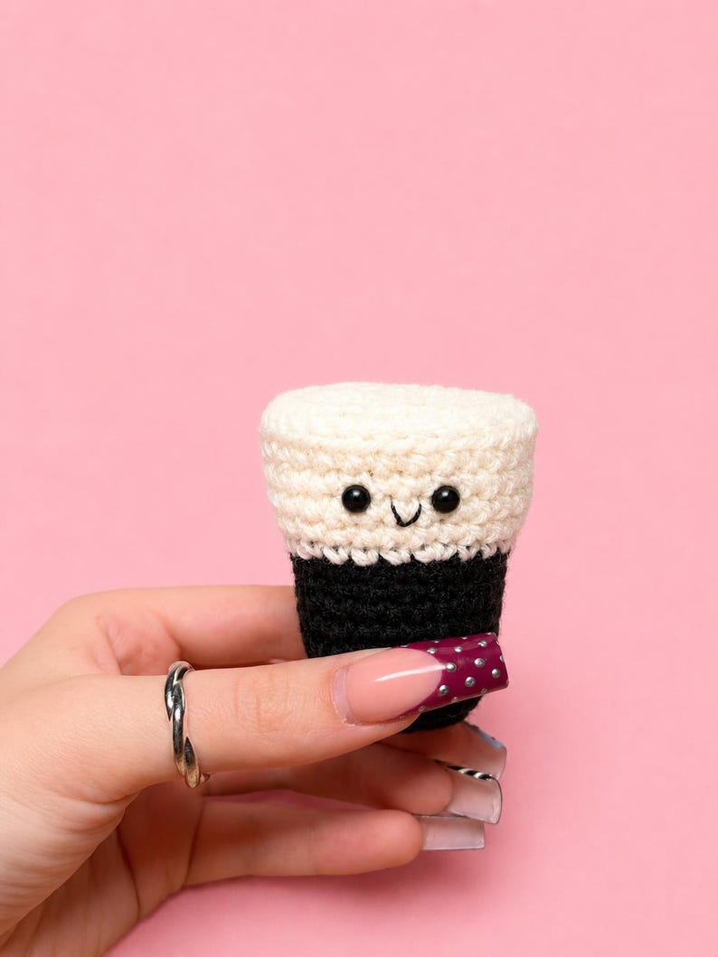 May include: A small, crocheted drink with a smiling face. The top is cream-colored, the middle is black, and the bottom is a dark burgundy with small white dots. The background is a solid pink.