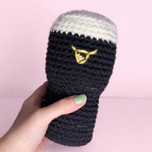 May include: A handmade crochet beer glass with a white top and dark gray body. A yellow embroidered design is on the front. The beer glass is held against a pink background.