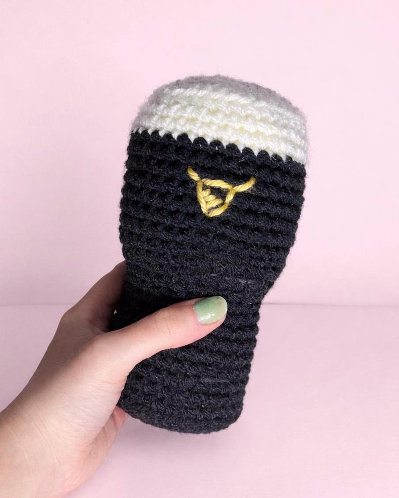 May include: A handmade crochet beer glass with a white top and dark gray body. A yellow embroidered design is on the front. The beer glass is held against a pink background.