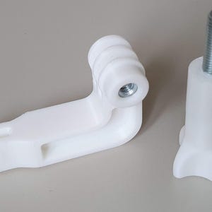 Può includere: Due componenti in plastica bianca, probabilmente per un sistema di montaggio. Uno è a forma di L con un inserto filettato, l'altro è un pezzo cilindrico con un bullone filettato. Entrambi sono su una superficie neutra.