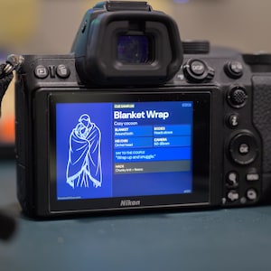 May include: A black Nikon digital camera with a screen displaying a blue graphic of a person wrapped in a blanket. The screen also shows text including "Blanket Wrap" and "Cozy cocoon". The camera has a black strap.