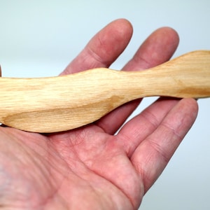 May include: A wooden butter knife with a smooth, light brown finish. The knife has a slightly curved handle and a rounded blade.