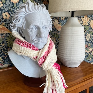 May include: A gray bust of Albert Einstein wearing a white and pink woven scarf with fringe. The scarf has a repeating pattern of pink stripes. The bust is on a wooden cabinet, with a lamp and floral wallpaper in the background.
