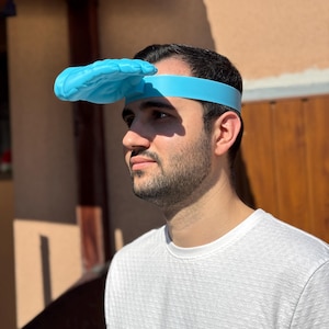 Könnte beinhalten: Ein neuartiges blaues Handvisier, das an einem blauen Stirnband befestigt ist. Die Hand befindet sich über der Stirn. Die Person auf dem Bild trägt ein weißes, strukturiertes T-Shirt.