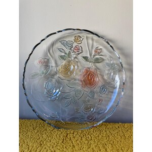 May include: A clear glass serving plate with a scalloped edge, featuring an embossed floral design of roses with subtle yellow, peach, and pink tinted petals and green leaves molded into the surface of the transparent glass material.