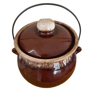 May include: A round ceramic pot with a glossy dark brown glaze and a textured, speckled beige rim. It features a matching lid with a knob handle, two side handles, and a thin, arched metal wire handle attached to the sides of the pot.