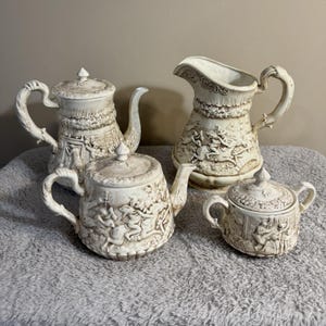 May include: A white ceramic mug with a black handle and the text "But First, Coffee" in a bold, black font. The mug is set against a blurred background, suggesting a cozy setting.