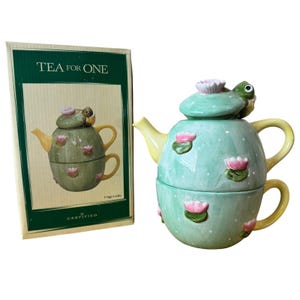 May include: A ceramic tea for one set featuring a light green teapot and cup stack decorated with white polka dots and pink water lily accents. The teapot lid is topped with a small green frog figure. The set includes a yellow handle and spout.