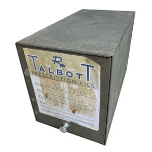 May include: A rectangular, olive-gray metal prescription file box with a small silver knob handle. A white paper label on the front features the text "TALBOTT PRESCRIPTION FILE" with a scale icon, handwritten numbers N69000 to N69999, and manufacturer details.