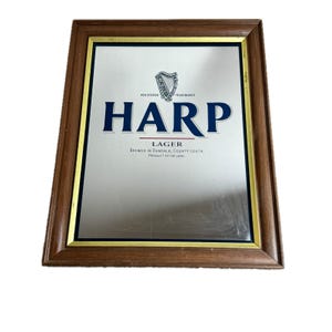 May include: A framed mirror with the Harp Lager logo. The mirror has a brown wooden frame with a gold inner border. The Harp logo is in blue, with the word "HARP" prominently displayed. The text "LAGER" and other details are also visible.
