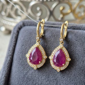 Puede incluir: Un par de pendientes colgantes dorados con gemas rojas en forma de pera. Los pendientes están rodeados de pequeñas gemas transparentes y presentan un diseño decorativo. Los pendientes se exhiben sobre un fondo gris.