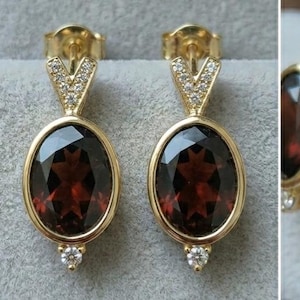 May include: A pair of gold earrings featuring oval, dark red gemstones. The earrings have a V-shaped top section and a small, round accent stone at the base. The gemstones are set in a gold frame, creating a classic and elegant appearance.