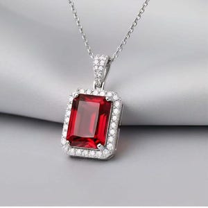 Peut inclure: Collier en argent avec un pendentif rectangulaire en pierre rouge foncé. Le pendentif est entouré d'un halo de petites pierres claires. La chaîne en argent délicate crée un look classique et élégant.
