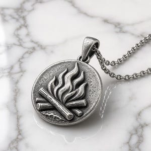 May include: A circular silver-toned pendant featuring a raised relief design of a campfire with logs and flames. The pendant has an antiqued finish and is attached to a matching silver-toned metal chain link necklace with a sturdy bail.